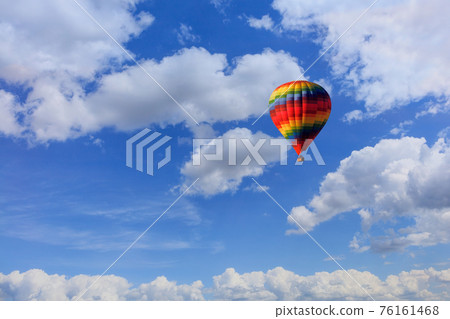A motley multicolored hot air balloon raises a basket with tourists in the blue sky among white clouds. 76161468