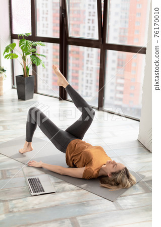 Slim adult caucasian woman does half-bridge pose on the floor in front of the laptop at home, online fitness class, vertical photo, selective focus. Slim adult caucasian woman does half-bridge pose on the floor in front of the laptop at home, online fitness class, vertical photo, selective focus. 76170010