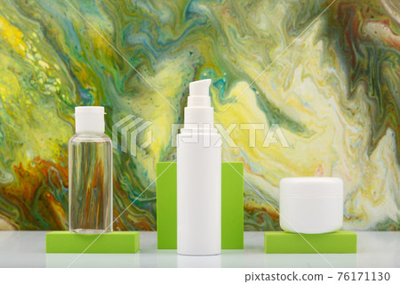 Skin lotion in transparent bottle, face cream and lip balm on green podiums against marbled background in green colors.  76171130