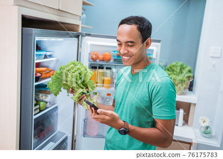 man standing in front of the fridge while holding his mobile phone 76175783