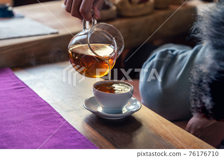Hand pouring hot tea from jug into white teacup on the table Hand pouring hot tea from jug into white teacup on the table 76176710