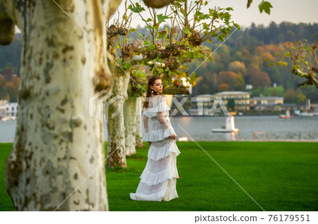 A bride in a white wedding dress in a park in an Austrian town with large trees at sunset A bride in a white wedding dress in a park in an Austrian town with large trees at sunset 76179551