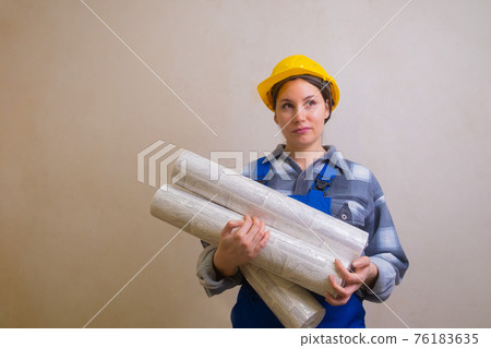 A girl in a blue construction worker's jumpsuit and a yellow hard hat holds wallpaper in her hands and looks thoughtfully up and to the right against the background of a gray stucco wall A girl in a blue construction worker's jumpsuit and a yellow hard hat holds wallpaper in her hands and looks thoughtfully up and to the right against the background of a gray stucco wall 76183635