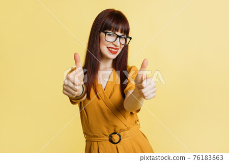Young caucasian businesswoman in trendy yellow dress and eye glasses, isolated on yellow background, giving a thumbs up gesture and smiling with teeth to camera 76183863