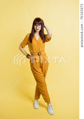 Full length view of young gorgeous red haired lady in bright yellow trendy overalls, touching hair, posing to camera with legs crossed on isolated yellow color background 76183873