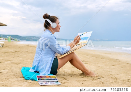 Mature woman sitting on sandy beach drawing sketch of sea Mature woman sitting on sandy beach drawing sketch of sea 76185258