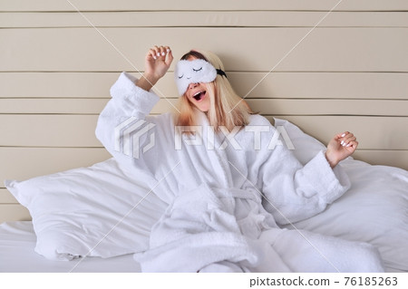 Young woman in white bed in bathrobe, eye mask, female yawns Young woman in white bed in bathrobe, eye mask, female yawns 76185263