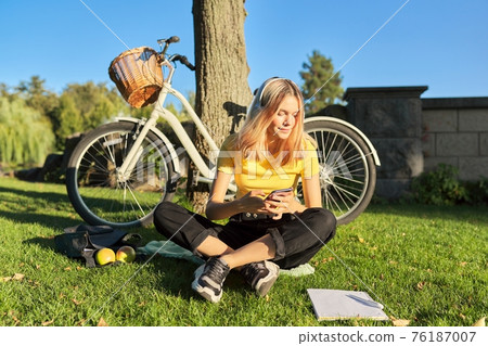 Teenage student in headphones with smartphone bicycle sitting in park Teenage student in headphones with smartphone bicycle sitting in park 76187007