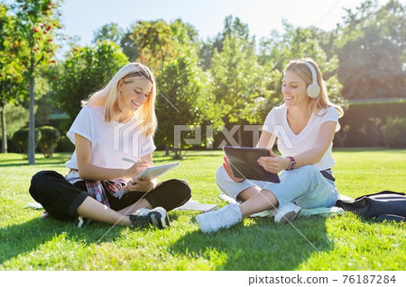 Girls teenage students sitting on green lawn in park with backpack, digital tablet Girls teenage students sitting on green lawn in park with backpack, digital tablet 76187284