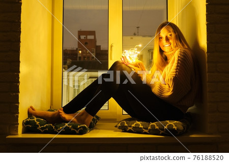 Winter portrait of woman sitting on windowsill, sparkling garland lights in hands of girl 76188520