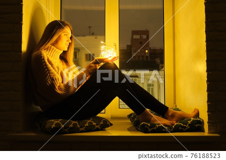Winter portrait of woman sitting on windowsill, sparkling garland lights in hands of girl Winter portrait of woman sitting on windowsill, sparkling garland lights in hands of girl 76188523