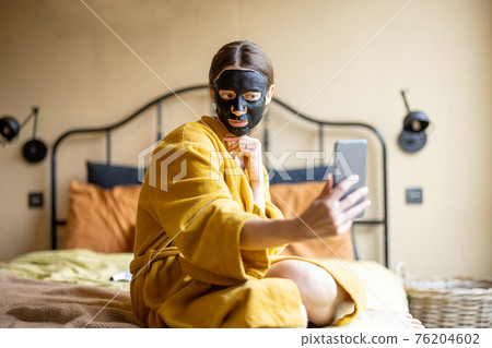 Woman in bathrobe with black cleansing mask and phone in bedroom 76204602