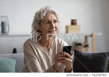 Happy pensive mature lady holding smartphone at home 76206768