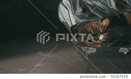 Forge workshop. Smithy. Worker in a welding hood helmet welds a part by electric welding. Sparks are reflected in the protective screen. Forge workshop. Smithy. Worker in a welding hood helmet welds a part by electric welding. Sparks are reflected in the protective screen. 76207839