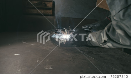 Forge workshop. Smithy. Worker in a welding hood helmet welds a part by electric welding. Sparks are reflected in the protective screen. Forge workshop. Smithy. Worker in a welding hood helmet welds a part by electric welding. Sparks are reflected in the protective screen. 76207840