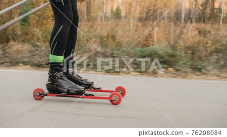 Training an athlete on the roller skaters. Biathlon ride on the roller skis with ski poles, in the helmet. Autumn workout. Roller sport. 76208084