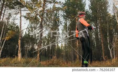 Training an athlete on the roller skaters. Biathlon ride on the roller skis with ski poles, in the helmet. Autumn workout. Roller sport. 76208107