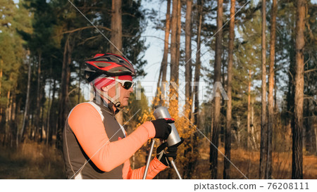 Training an athlete on the roller skaters. Biathlon ride on the roller skis with ski poles, in the helmet. Autumn workout. Roller sport. Athlete drinks water from flask. Training an athlete on the roller skaters. Biathlon ride on the roller skis with ski poles, in the helmet. Autumn workout. Roller sport. Athlete drinks water from flask. 76208111