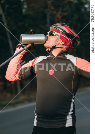 Training an athlete on the roller skaters. Biathlon ride on the roller skis with ski poles, in the helmet. Autumn workout. Roller sport. Athlete drinks water from flask. 76208773