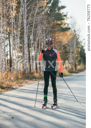 Training an athlete on the roller skaters. Biathlon ride on the roller skis with ski poles, in the helmet. Autumn workout. Roller sport. 76208775