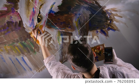 Artist designer draws an eagle on wall. Craftsman decorator paints picture with acrylic oil color looking at sketch in phone. Dark magic cinematic. Artist designer draws an eagle on wall. Craftsman decorator paints picture with acrylic oil color looking at sketch in phone. Dark magic cinematic. 76208891