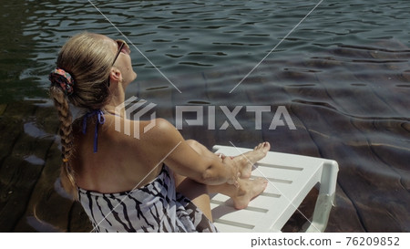 Woman lie on a sunbed in sunglasses and a boho silk shawl. Girl rest on a flood wood underwater pier. The pavement is covered with water in the lake. 76209852