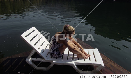 Woman lie on a sunbed in sunglasses and a boho silk shawl. Girl rest on a flood wood underwater pier. The pavement is covered with water in the lake. 76209881