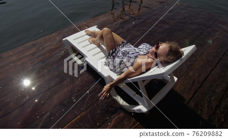 Woman lie on a sunbed in sunglasses and a boho silk shawl. Girl rest on a flood wood underwater pier. The pavement is covered with water in the lake. 76209882