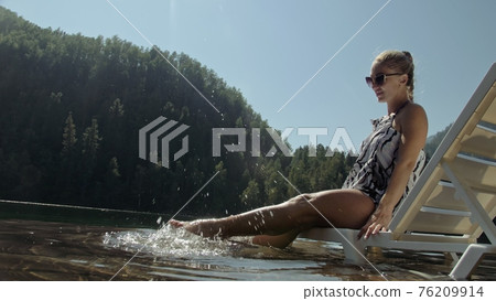 Woman lie on a sunbed in sunglasses and a boho silk shawl. Girl rest on a flood wood underwater pier. The pavement is covered with water in the lake. 76209914