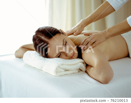 Pretty brunette woman enjoying procedure of back massage in spa salon. Beauty concept 76210013