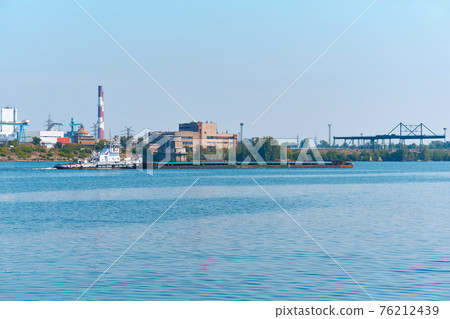 towboat pushes dry bulk cargo barge on the river against the backdrop of an industrial landscape on the shore 76212439