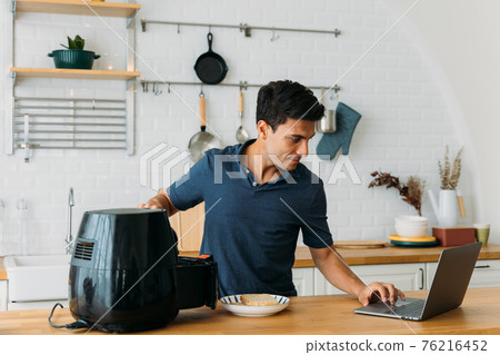 Man using air fryer and laptop in kitchen Man using air fryer and laptop in kitchen 76216452
