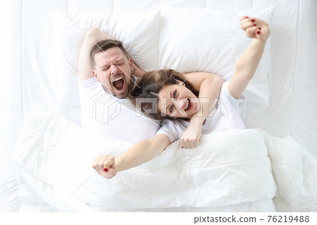 Yawning happy man and woman lie on large white bed 76219488