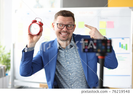 Smiling businessman holds red alarm clock and conducts online time management courses Smiling businessman holds red alarm clock and conducts online time management courses 76219548