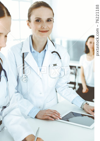 Two woman-doctors at work with patient at background. Female physicians filling up medical documents or prescription while standing in hospital reception desk. Data in medicine Two woman-doctors at work with patient at background. Female physicians filling up medical documents or prescription while standing in hospital reception desk. Data in medicine 76221188