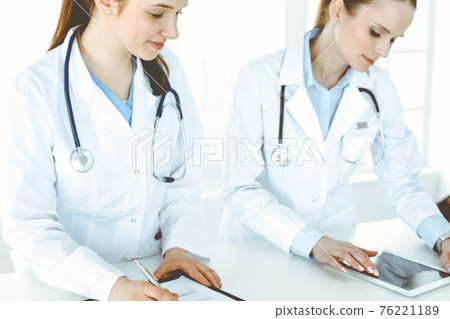 Two woman-doctors at work with patient at background. Female physicians filling up medical documents or prescription while standing in hospital reception desk. Data in medicine Two woman-doctors at work with patient at background. Female physicians filling up medical documents or prescription while standing in hospital reception desk. Data in medicine 76221189