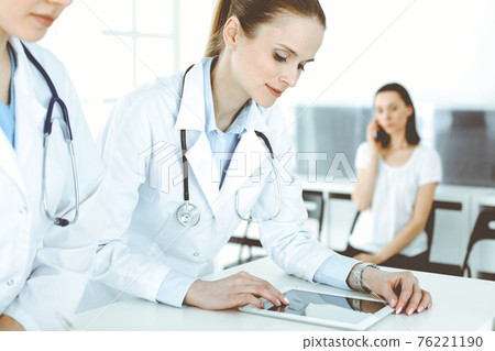 Two woman-doctors at work with patient at background. Female physicians filling up medical documents or prescription while standing in hospital reception desk. Data in medicine Two woman-doctors at work with patient at background. Female physicians filling up medical documents or prescription while standing in hospital reception desk. Data in medicine 76221190