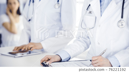Unknown woman-doctors at work with patient at the background. Female physicians filling up medical documents or prescription while standing in hospital reception desk, close-up. Health care concept 76221192
