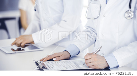 Unknown woman-doctors at work with patient at the background. Female physicians filling up medical documents or prescription while standing in hospital reception desk, close-up. Health care concept Unknown woman-doctors at work with patient at the background. Female physicians filling up medical documents or prescription while standing in hospital reception desk, close-up. Health care concept 76221193