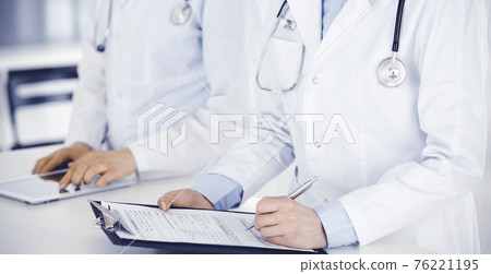 Unknown woman-doctors at work with patient at the background. Female physicians filling up medical documents or prescription while standing in hospital reception desk, close-up. Health care concept Unknown woman-doctors at work with patient at the background. Female physicians filling up medical documents or prescription while standing in hospital reception desk, close-up. Health care concept 76221195