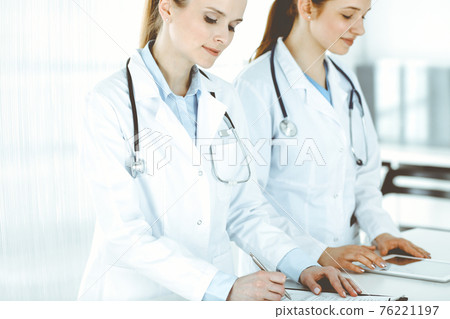 Woman-doctor cheerful smiling at work with colleague and patient at background. Portrait of middle aged female physician filling up medical documents or prescription while standing in hospital Woman-doctor cheerful smiling at work with colleague and patient at background. Portrait of middle aged female physician filling up medical documents or prescription while standing in hospital 76221197