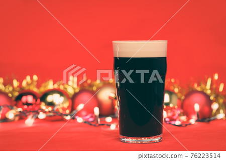 Dark ale beer in pint glass with christmas baubles tinsel and lights on red background toned Dark ale beer in pint glass with christmas baubles tinsel and lights on red background toned 76223514