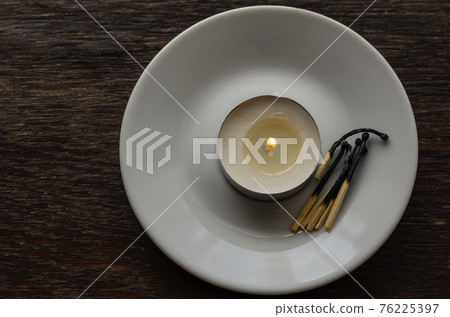 Burning candle in a white saucer on a wooden table. Burning candle in a white saucer on a wooden table. 76225397