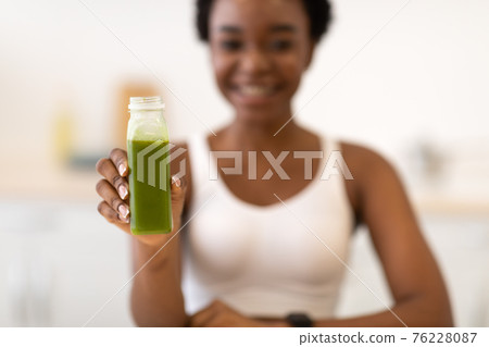 Slim African Female Showing Green Smoothie Bottle Sitting In Kitchen Slim African Female Showing Green Smoothie Bottle Sitting In Kitchen 76228087