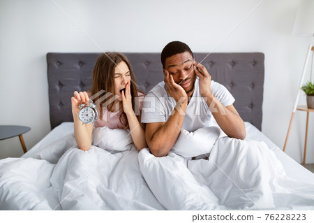 Interracial married couple having problem with waking up in morning, sitting sleepy in bed with alarm clock Interracial married couple having problem with waking up in morning, sitting sleepy in bed with alarm clock 76228223