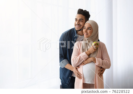 Portrait Of Happy Pregnant Islamic Couple Standing Near Window At Home 76228522