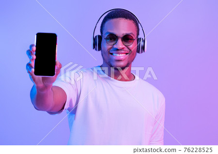 Joyful Young Black Man In Wireless Headphones Showing Cellphone With Blank Screen 76228525