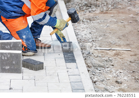 A worker hammers in the paving slabs of the work site with a rubber hammer. A worker hammers in the paving slabs of the work site with a rubber hammer. 76228551