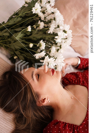 A beautiful brunette in a red dress lies on the bed next to a bouquet of daisies 76229461