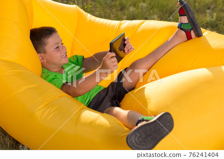 cheerful boy lies in a yellow lamzack, holding a tablet in his hands, plays or communicates on social networks, concept cheerful boy lies in a yellow lamzack, holding a tablet in his hands, plays or communicates on social networks, concept 76241404